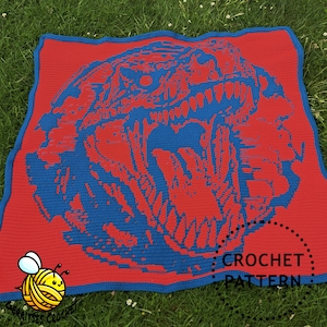 Op de afbeelding: Een rood gehaakt deken met een blauw dinosauruskopontwerp. De bek van de dinosaurus staat open en zijn tanden zijn zichtbaar. Het deken heeft een gehaakte rand en de tekst "CROCHET PATTERN" is zichtbaar in de rechterbenedenhoek.