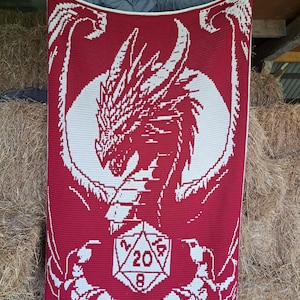 Puede incluir: Una manta de crochet roja con un dise&ntilde;o de drag&oacute;n blanco que sostiene un dado de 20 caras. La manta tiene un borde blanco.