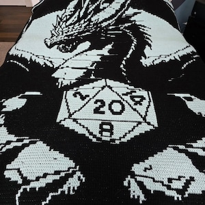 Puede incluir: Manta de crochet en blanco y negro con un drag&oacute;n con un dado de 20 caras en el centro. La manta tiene un dise&ntilde;o blanco y azul claro.