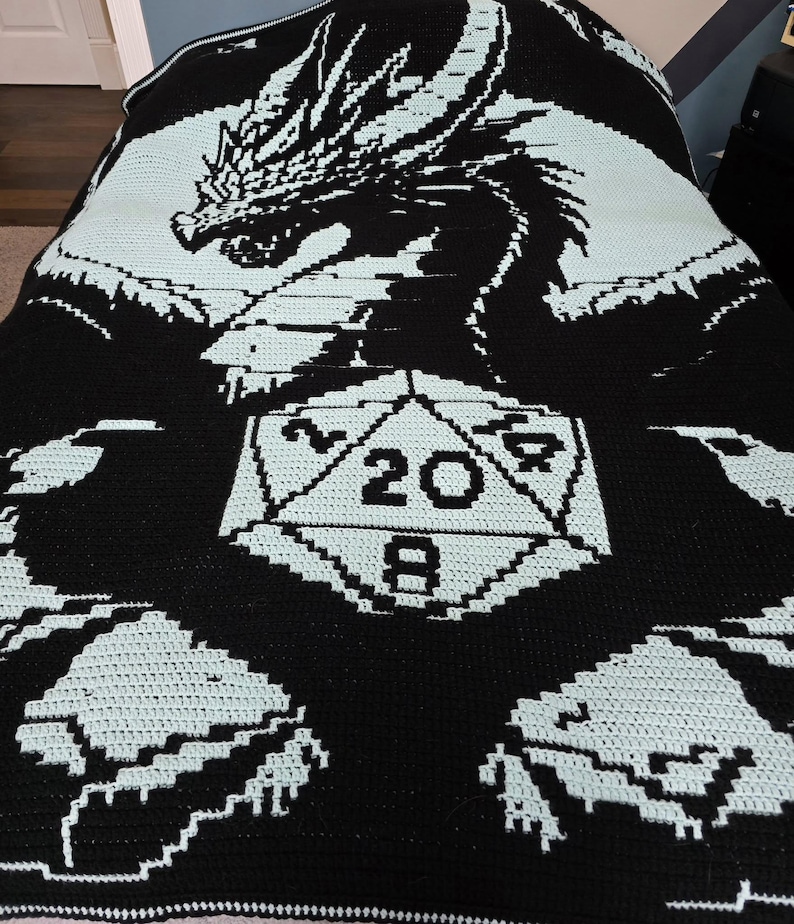 Puede incluir: Manta de crochet en blanco y negro con un drag&oacute;n con un dado de 20 caras en el centro. La manta tiene un dise&ntilde;o blanco y azul claro.