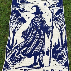 May include: A crocheted blanket featuring a wizard in a dark blue and white design. The wizard is depicted with a staff, hat, and cloak, set against a forest and starry sky backdrop. The blanket is rectangular with a white border.