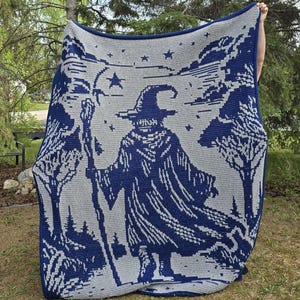 May include: A large, crocheted blanket featuring a wizard design in shades of blue and gray. The wizard is holding a staff and is surrounded by trees and stars. The blanket is held up outdoors.