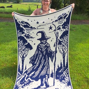 May include: A large, cream-colored blanket with a dark blue wizard design. The blanket features a wizard holding a staff, surrounded by trees and stars. The design is a pixelated style, with a white border.