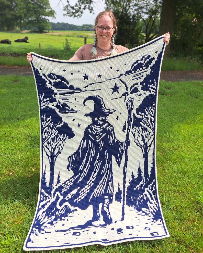 May include: A large, cream-colored blanket with a dark blue wizard design. The blanket features a wizard holding a staff, surrounded by trees and stars. The design is a pixelated style, with a white border.