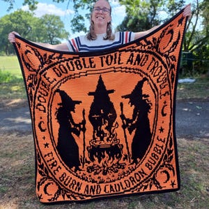 May include: A rectangular crochet blanket featuring a vibrant orange background with black details. The design displays two silhouettes of witches standing on either side of a bubbling cauldron over a fire, with the text "DOUBLE, DOUBLE TOIL AND TROUBLE, FIRE BURN AND CAULDRON BUBBLE" encircling the central image.