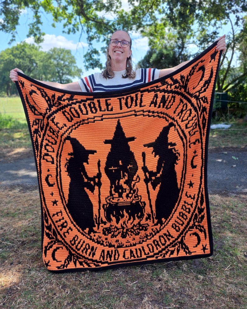 May include: A rectangular crochet blanket featuring a vibrant orange background with black details. The design displays two silhouettes of witches standing on either side of a bubbling cauldron over a fire, with the text "DOUBLE, DOUBLE TOIL AND TROUBLE, FIRE BURN AND CAULDRON BUBBLE" encircling the central image.