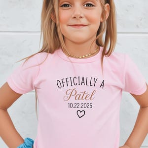 Könnte beinhalten: Hellrosa T-Shirt mit dem Schriftzug "Officially a Patel" in Schreibschrift, dem Datum "10.22.2025" und einem Herz. Das Shirt wird von einem jungen Mädchen mit blonden Haaren getragen.