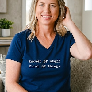 Könnte beinhalten: Ein marineblaues V-Ausschnitt-T-Shirt mit dem weißen Text "knower of stuff fixer of things". Die Person trägt eine blaue Jeans und sitzt auf einem grauen Sessel. Der Hintergrund umfasst ein Fenster und eine Lampe.
