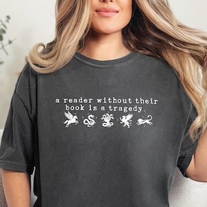 Könnte beinhalten: Dunkelgraues T-Shirt mit dem Text "a reader without their book is a tragedy." Das Shirt zeigt weiße Illustrationen eines Pegasus, einer Schlange, eines Greifs und eines Löwen. Das Shirt ist aus weichem Material.