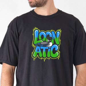 Könnte beinhalten: Ein schwarzes T-Shirt mit kurzen Ärmeln und einem zentralen Aufdruck mit dem Text "LOON ATIC" in blauer und grüner Graffiti-Schrift. Eine detaillierte Abbildung eines Eistauchers befindet sich zwischen den Wörtern auf einem hellgrünen Hintergrund.
