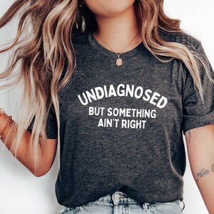Könnte beinhalten: Ein dunkel anthrazitfarbenes Kurzarm-T-Shirt mit dem weißen Text UNDIAGNOSED BUT SOMETHING AIN'T RIGHT auf der Brust. Das T-Shirt wird mit einer hellblauen Jeans mit ausgefranstem Saum und einem einzelnen weißen Slipper kombiniert.