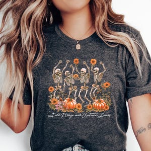 May include: Dark gray t-shirt featuring a graphic of dancing skeletons holding sunflowers, with pumpkins and flowers. The text "Fall Breeze and Autumn Leaves" is printed below the graphic. The shirt is paired with light blue jeans.