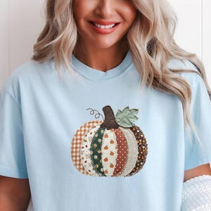 May include: A light blue short-sleeve t-shirt featuring a central graphic of a pumpkin made from various fabric patches. The pumpkin design includes patterns of gingham, floral prints, polka dots, and small pumpkin motifs in brown, green, and cream.