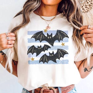 Könnte beinhalten: Ein cremefarbenes T-Shirt mit einem Grafikdruck von drei schwarzen Fledermäusen, die über horizontale, hellblaue, abgenutzte Streifen fliegen. Das Design ist mit kleinen goldfarbenen Sternen akzentuiert, die zwischen den Fledermäusen verteilt sind.