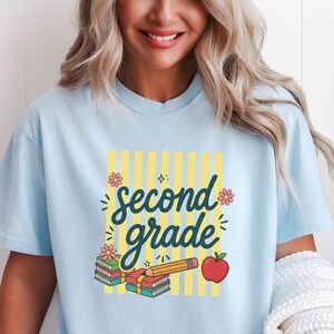 Könnte beinhalten: Ein hellblaues T-Shirt mit einem Aufdruck, der den Text second grade in dunkelblauer Schreibschrift zeigt. Das Design umfasst gelbe vertikale Streifen, einen Bücherstapel, einen gelben Bleistift, einen roten Apfel und kleine rosa Blumen.