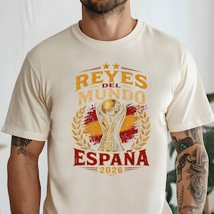 Könnte beinhalten: Ein cremefarbenes Kurzarm-T-Shirt mit einem Grafikdruck, der den Text REYES DEL MUNDO ESPAÑA 2026 zeigt. Das Design umfasst eine zentrale Trophäenillustration, einen stilisierten spanischen Flaggenhintergrund und goldene Lorbeerblattakzente.