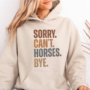 Könnte beinhalten: Beigefarbener Kapuzenpullover mit dem Text "SORRY. CAN'T. HORSES. BYE." in braunen, grauen und beigen Buchstaben. Der Hoodie hat eine Vordertasche und eine Kapuze. Die Person trägt hellblaue Jeans mit zerrissenen Knien.