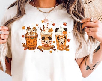 Può includere: T-shirt color crema con tre tazze di caffè a fumetti con disegni a tema autunnale. Le tazze raffigurano Cip e Ciop in una zucca, Topolino e Minnie, circondati da foglie autunnali e bastoncini di cannella. La maglietta è abbinata a pantaloncini di jeans blu e una cintura marrone.