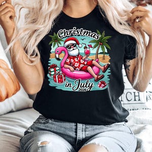 May include: A black t-shirt featuring a graphic of Santa Claus wearing sunglasses and a red floral shirt, relaxing in a pink flamingo pool float on a beach. The design includes palm trees, wrapped gifts, and the text Christmas in July 2026.
