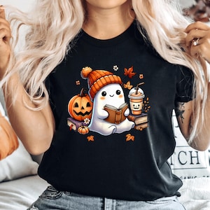 Könnte beinhalten: Schwarzes T-Shirt mit einem Cartoon-Geist, der eine orangefarbene Mütze trägt und ein Buch liest. Ein Kürbis, eine Kaffeetasse und Herbstblätter sind ebenfalls abgebildet. Das Design hat ein Herbst- oder Halloween-Thema.