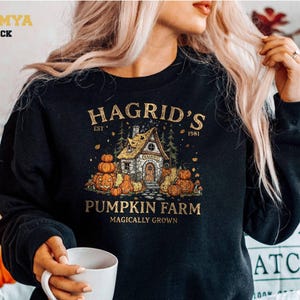 Könnte beinhalten: Ein schwarzes Langarm-Sweatshirt mit einem Grafikdruck, der ein Steinhäuschen umgeben von Kürbissen und Bäumen zeigt. Der Text lautet HAGRID'S EST 1981 PUMPKIN FARM MAGICALLY GROWN. Das Design verwendet orange, gelbe und braune Töne auf dem schwarzen Stoff.