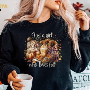 Könnte beinhalten: Ein schwarzes Langarm-Sweatshirt mit einem Grafikdruck und dem Text "Just a girl who loves fall". Das Design zeigt eine Strickmütze, einen Kürbis, eine Sonnenblume, einen Kaffeebecher und einen Football, umgeben von herbstlichen Blättern.