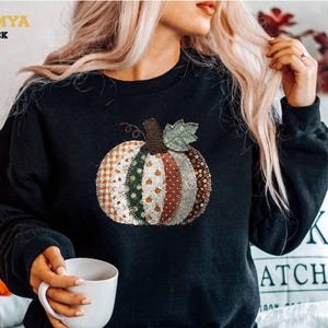 Puede incluir: Una sudadera negra de cuello redondo con un gráfico central de una calabaza compuesta por varios segmentos de tela estampada, incluyendo cuadros vichy naranjas, motivos florales verdes y lunares, con un tallo marrón y una hoja verde.