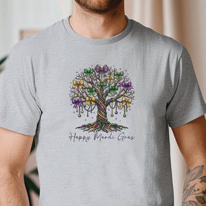 Könnte beinhalten: Hellgraues T-Shirt mit einem farbenfrohen Baumdesign mit lila, grünen und goldenen Akzenten. Der Baum ist mit Schleifen und Perlen verziert, darunter der Text "Happy Mardi Gras". Das T-Shirt hat kurze Ärmel und ist für den Alltag geeignet.