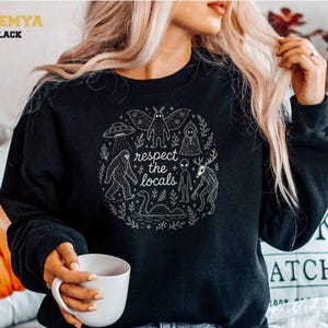 Peut inclure: Un sweat-shirt noir à manches longues avec une impression graphique circulaire blanche portant le texte "respect the locals" entouré d'illustrations d'un homme-phalène, d'un OVNI, du Bigfoot, d'une silhouette encapuchonnée, d'un alien et d'un monstre.
