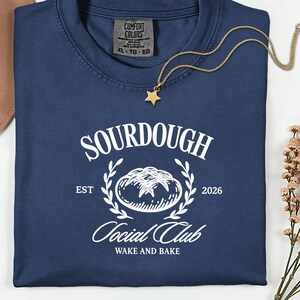 Könnte beinhalten: Marineblaues T-Shirt mit weißem Aufdruck "SOURDOUGH Social Club". Das Design zeigt eine Brotillustration und den Text "WAKE AND BAKE". Eine goldene Stern-Anhänger-Halskette und ein brauner Hut sind ebenfalls sichtbar.