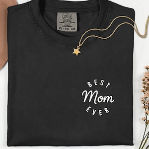 Könnte beinhalten: Schwarzes T-Shirt mit Rundhalsausschnitt und dem weißen Text "Best Mom Ever" in einem kreisförmigen Design. Eine goldene Halskette mit Sternanhänger liegt auf dem Shirt. Ein brauner Hut ist oben links zu sehen.