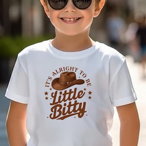 Peut inclure: T-shirt blanc avec un motif de chapeau de cowboy marron et le texte "It's Alright to be Little Bitty". Le t-shirt à manches courtes est porté par un enfant portant des lunettes de soleil.