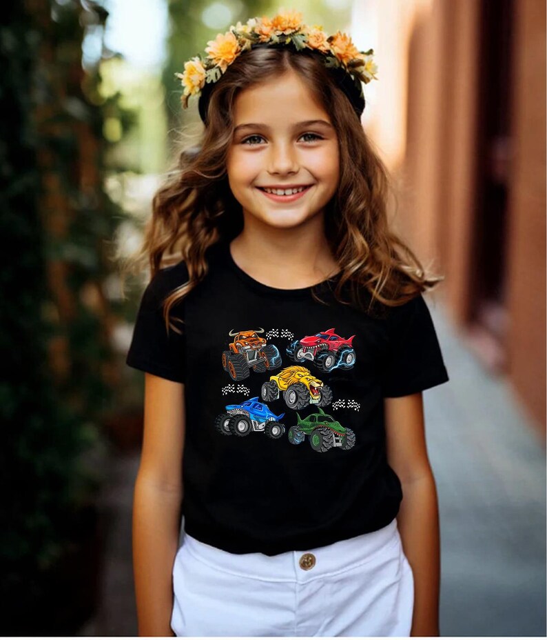 Puede incluir: Camiseta negra con dise&ntilde;os de monster trucks coloridos. Los camiones son de varios colores, incluyendo naranja, rojo, amarillo, azul y verde. La camiseta la lleva una persona con una corona de flores.