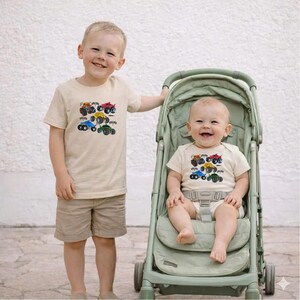 Puede incluir: Dos ni&ntilde;os con camisetas beige a juego con gr&aacute;ficos de monster trucks coloridos. Un ni&ntilde;o est&aacute; de pie, el otro est&aacute; sentado en un cochecito verde. Las camisetas tienen cuello redondo y mangas cortas. El ni&ntilde;o de pie lleva pantalones cortos color caqui.