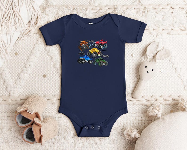 Puede incluir: Body de beb&eacute; azul marino con un gr&aacute;fico colorido de cinco monster trucks. Los camiones son rojos, amarillos, azules, verdes y naranjas, cada uno con dise&ntilde;os &uacute;nicos. El body tiene mangas cortas y cierres de presi&oacute;n.