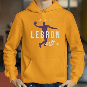 Könnte beinhalten: Senfgelber Kapuzenpullover mit Fronttasche. Der Hoodie zeigt eine lila Silhouette eines Basketballspielers mit dem Wort "LEBRON" in weißen Blockbuchstaben. Über dem Spieler steht das Wort "AIR".
