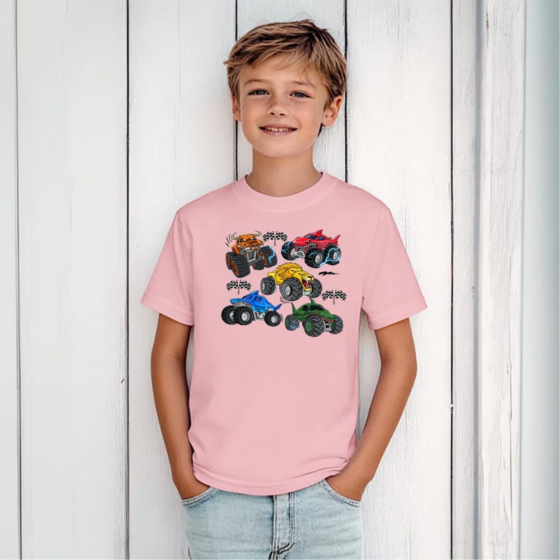 Puede incluir: Una camiseta rosa claro con un gr&aacute;fico colorido de seis monster trucks. Los camiones son de varios colores, incluyendo naranja, rojo, amarillo, azul y verde, con neum&aacute;ticos negros y banderas a cuadros. La camiseta la lleva una persona joven.
