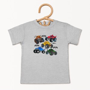 Puede incluir: Una camiseta gris claro con un gr&aacute;fico colorido de cuatro monster trucks. Los camiones son de color naranja, rojo, amarillo, azul y verde, con neum&aacute;ticos negros y banderas a cuadros. La camiseta est&aacute; colgada en una percha de madera.