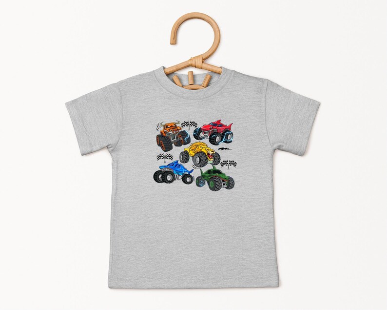 Puede incluir: Una camiseta gris claro con un gr&aacute;fico colorido de cuatro monster trucks. Los camiones son de color naranja, rojo, amarillo, azul y verde, con neum&aacute;ticos negros y banderas a cuadros. La camiseta est&aacute; colgada en una percha de madera.