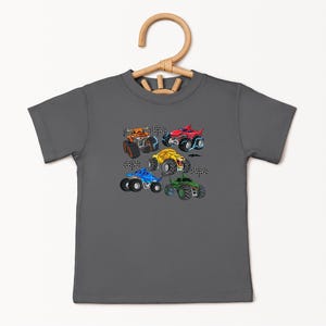 Puede incluir: Una camiseta gris carb&oacute;n con un gr&aacute;fico colorido de cinco monster trucks. Los camiones son de varios colores, incluyendo naranja, rojo, amarillo, azul y verde, con dise&ntilde;os detallados y banderas a cuadros.