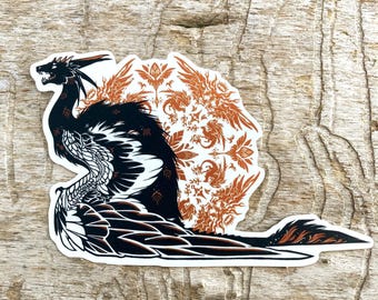 May include: Black and orange sticker featuring a stylized dragon with intricate details. The dragon is surrounded by a circular pattern of smaller dragons and floral motifs.