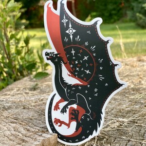 May include: Black and red dragon sticker with a white background. The dragon has a large wing with a geometric pattern and a small wing with a red and white pattern. The dragon is curled up in a defensive position.