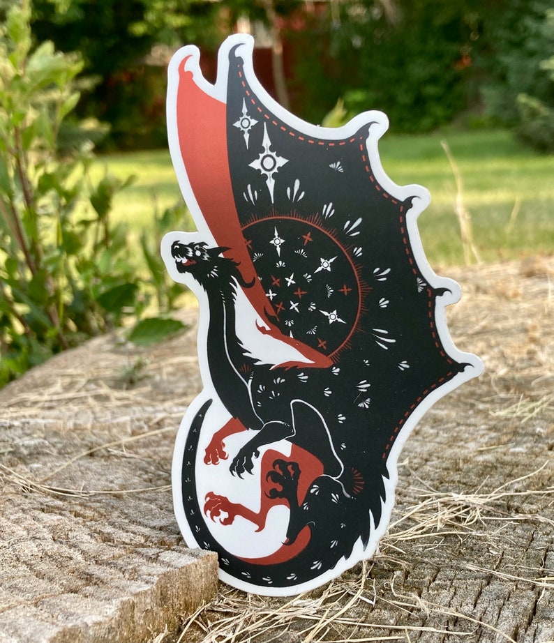 May include: Black and red dragon sticker with a white background. The dragon has a large wing with a geometric pattern and a small wing with a red and white pattern. The dragon is curled up in a defensive position.