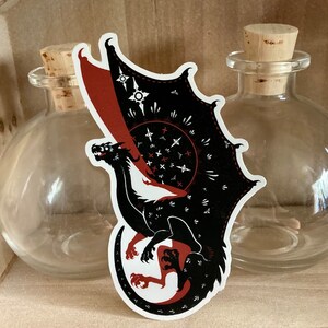 May include: Black and red dragon sticker with a white background. The dragon is curled up with its wings spread out. The dragon has a red and black patterned design on its wings.