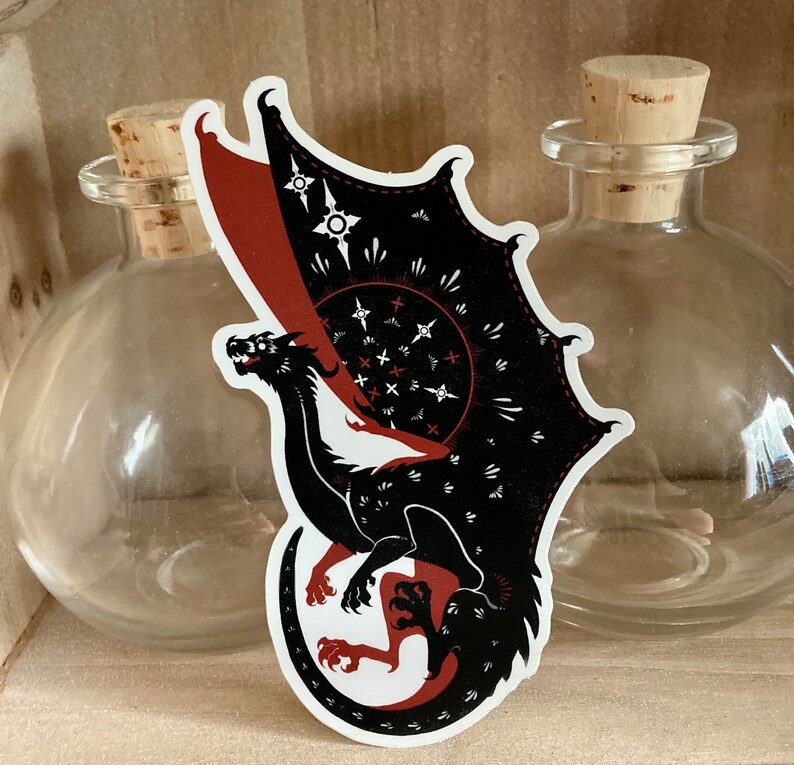May include: Black and red dragon sticker with a white background. The dragon is curled up with its wings spread out. The dragon has a red and black patterned design on its wings.