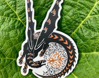 May include: A black and orange dragon sticker with intricate wing and tail details. The dragon is set against a white background with a detailed, swirling design. The sticker is placed on a large, green leaf.