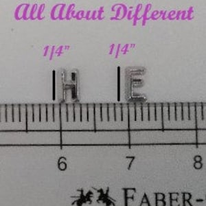 May include: Two silver metal letter beads, each measuring 1/4 inch tall. The letters are "H" and "E". The beads are shown on a ruler, with the "H" bead at the 6-inch mark and the "E" bead at the 7-inch mark.