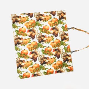 May include: A white tote bag featuring a repeating pattern of turkeys, pumpkins, and fall foliage. The turkeys are brown with red wattles, and the pumpkins are orange. The bag has a matching strap.