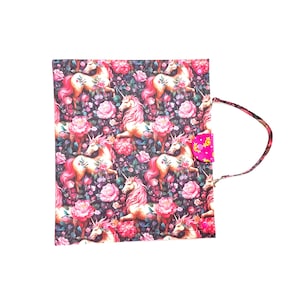 May include: A square, floral-patterned bag featuring unicorns and pink roses on a dark background. The bag has a pink button closure and a matching strap. The overall design is whimsical and colorful.