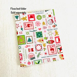 May include: A rectangular Christmas-themed floss bed folder with a white background. The folder is covered in colorful postage stamp designs featuring Christmas trees, gingerbread men, and snowflakes. The text "A QUIET LITTLE CHRISTMAS" is visible.